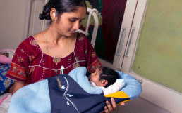 Woman holding a baby covered with a The Embrace Care, an infant warmer.