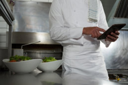 Mid-section of male chef using mobile phone in the kitchen for cutting hospitality food waste.